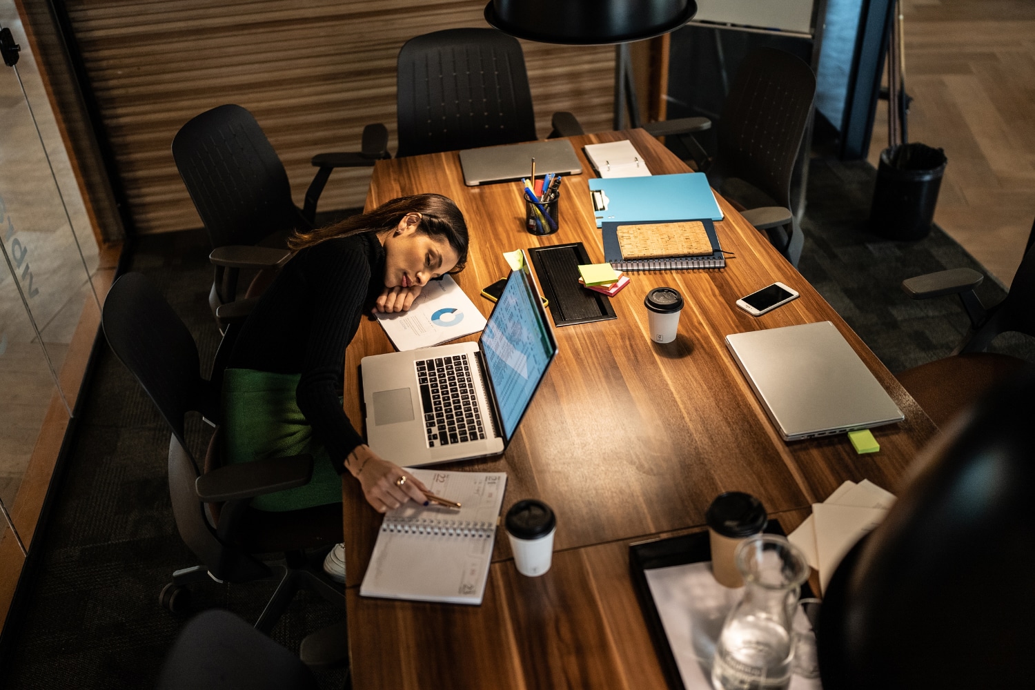 Một phụ nữ đang ngủ gục trên bàn làm việc với máy tính mở và nhiều đồ dùng xung quanh